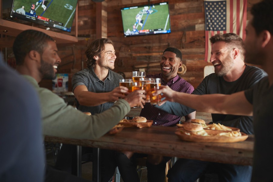 Five people at a restaurant table clinking glasses of beer. On the wall are TVs displaying football games.