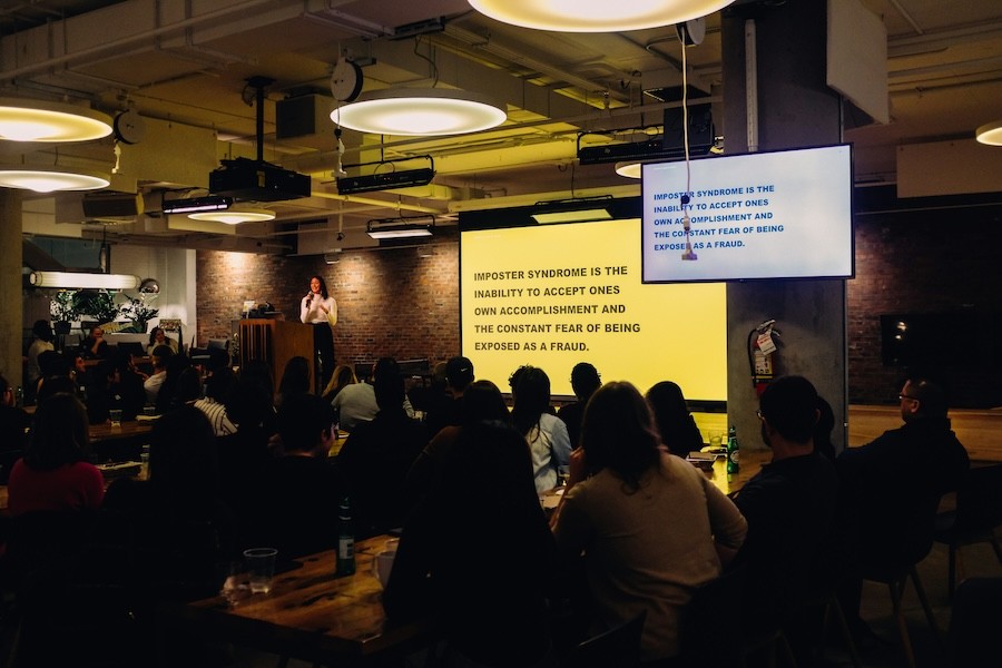 woman giving a presentation to a large corporate audience. Large screens show slide decks.