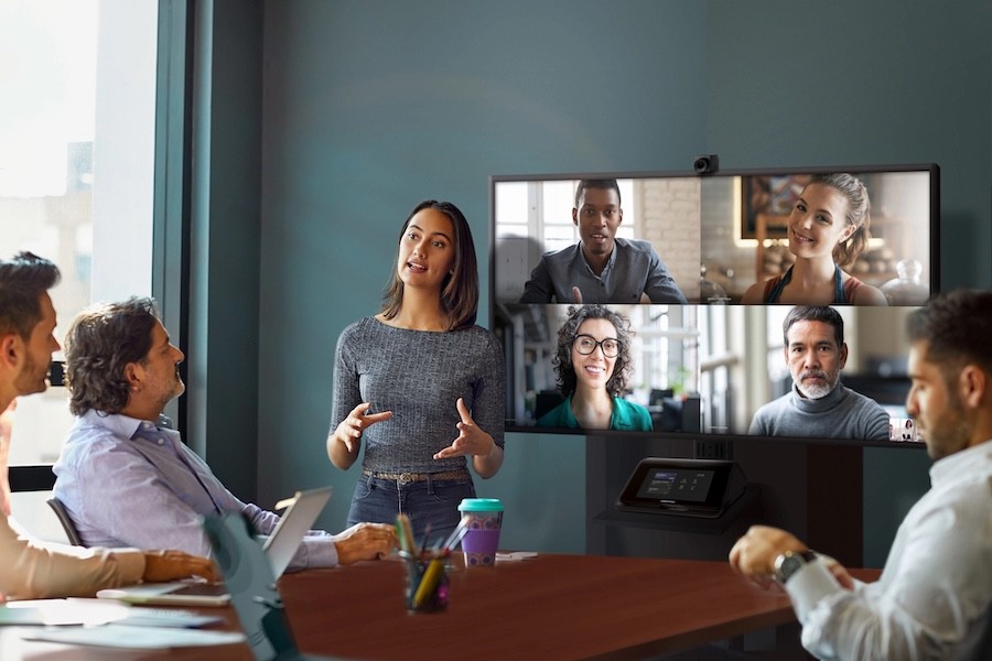 A woman gives a presentation in a meeting room while part of the team attends via video conference.