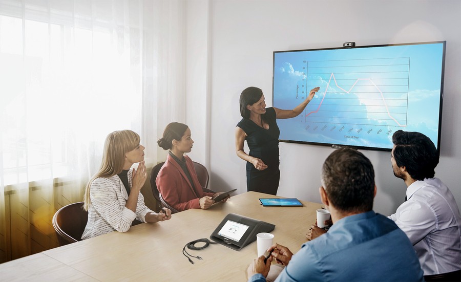 A team is holding a meeting in a conference room with a presenter motioning to a chart on the display in the front of the room.