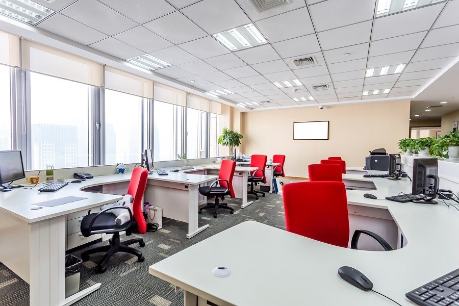 Modern open-plan office with red chairs, white desks, large windows, and motorized roller shades partially drawn to reduce glare.