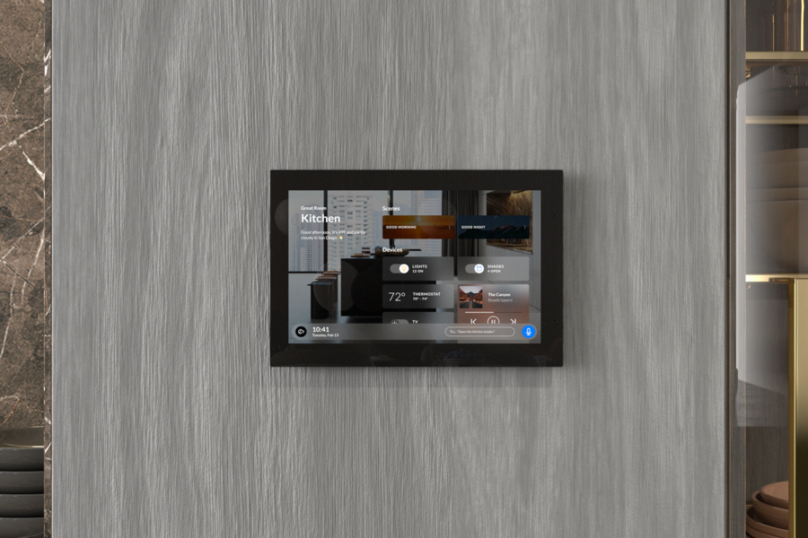 Wall-mounted smart home control panel in a modern kitchen, displaying lighting, thermostat, shades, and music controls.