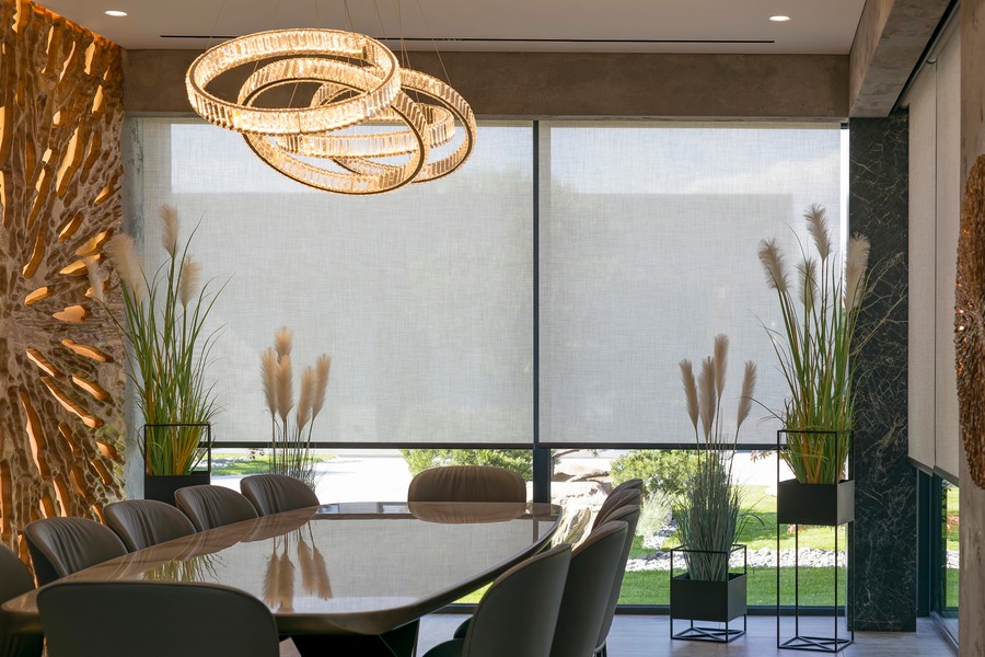 Modern conference room with motorized window shades, decorative lighting, and tall plants by large windows.
